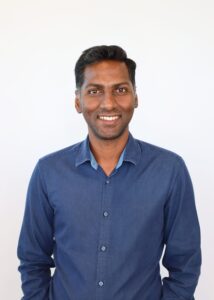 Karthik Dega smiling while wearing a blue button down shirt in front of white background